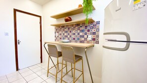 A compact kitchen with a white refrigerator, two bar stools, a small table, and a decorative tile backsplash. - Apartment with Balcony and Parking near Porto da Barra Beach (Salvador)