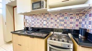 A compact kitchen with a gas stove, a microwave, and a tiled backsplash. - Apartment with Balcony and Parking near Porto da Barra Beach (Salvador)
