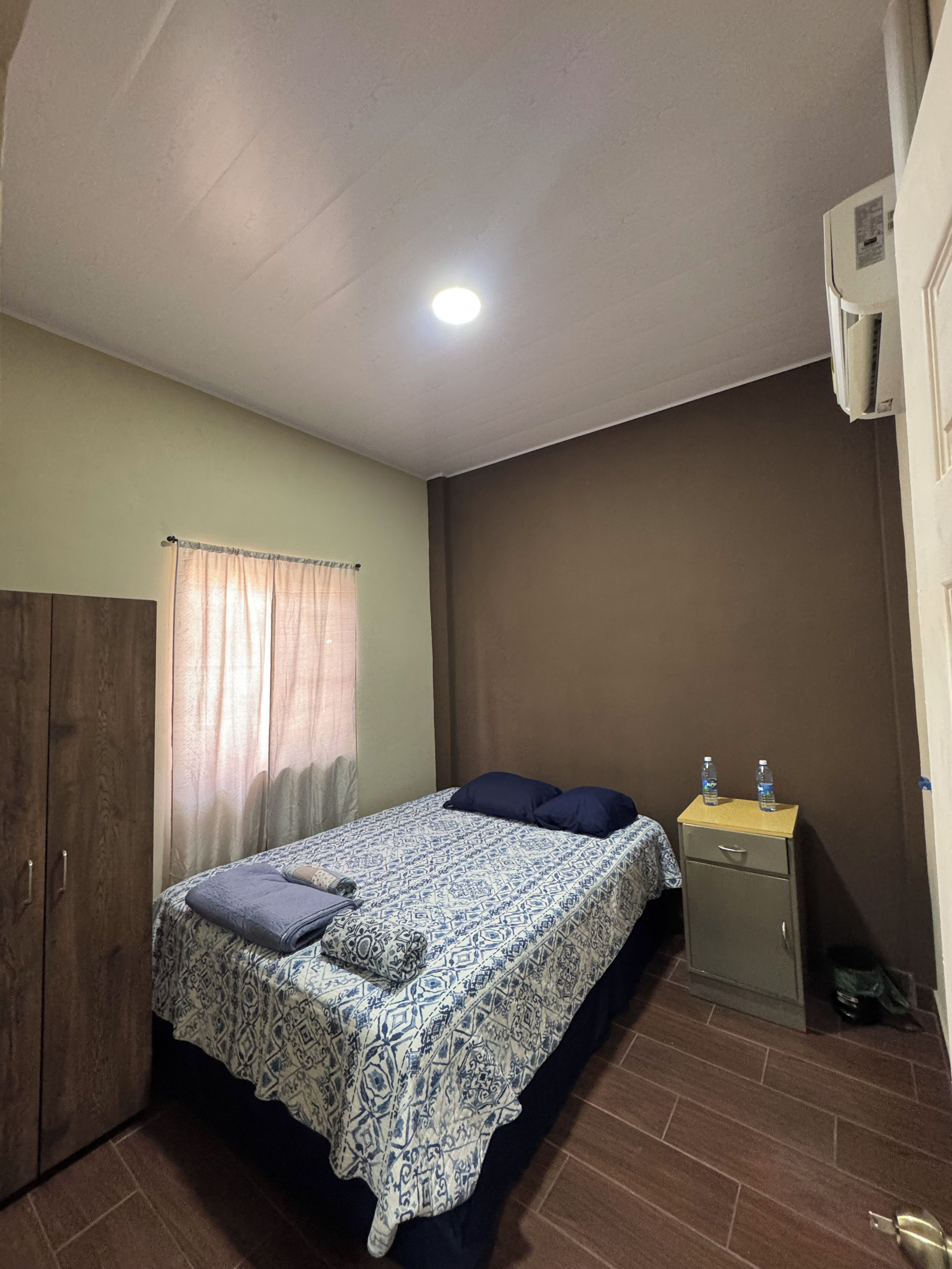 Une chambre à coucher comprenant un lit, une armoire, une table de chevet avec des bouteilles d’eau et un luminaire au plafond.
