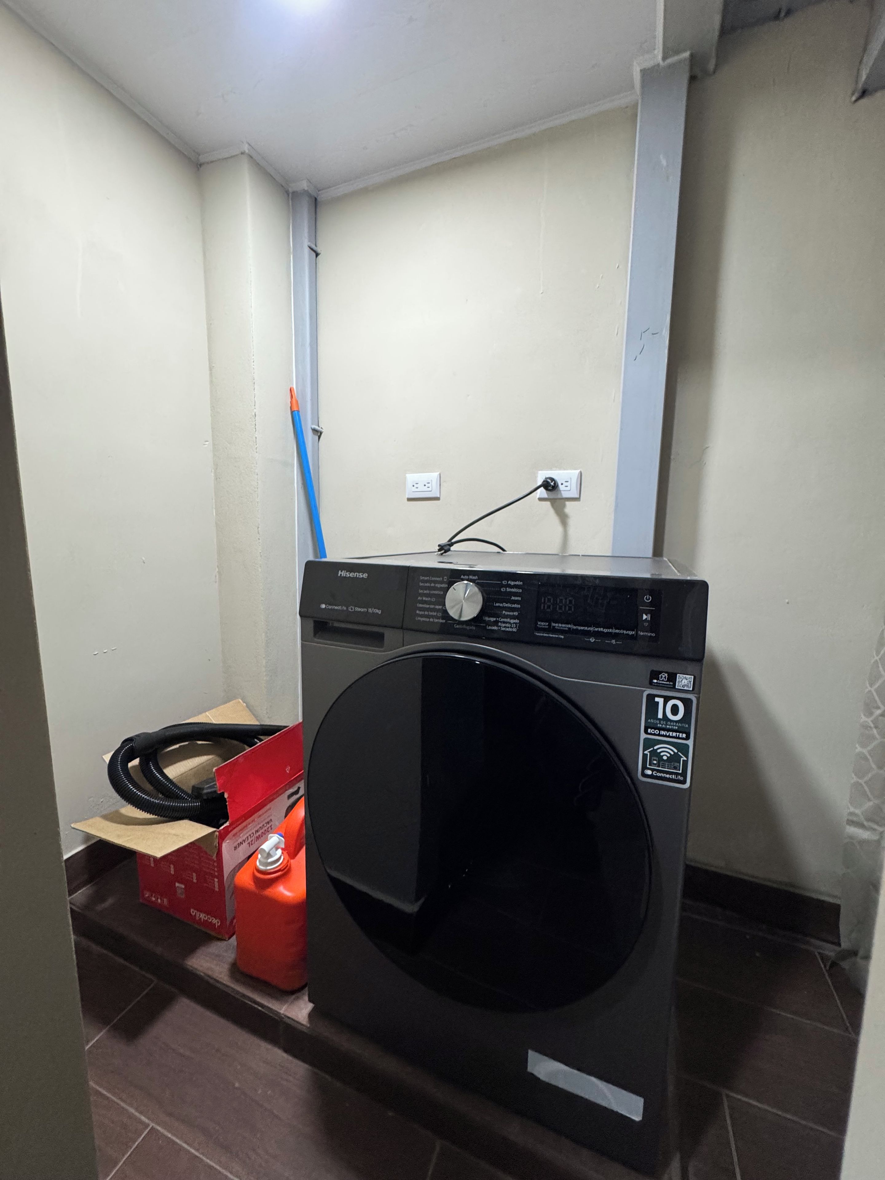 A compact washing machine with a control panel, a power outlet, and a red box on the floor.