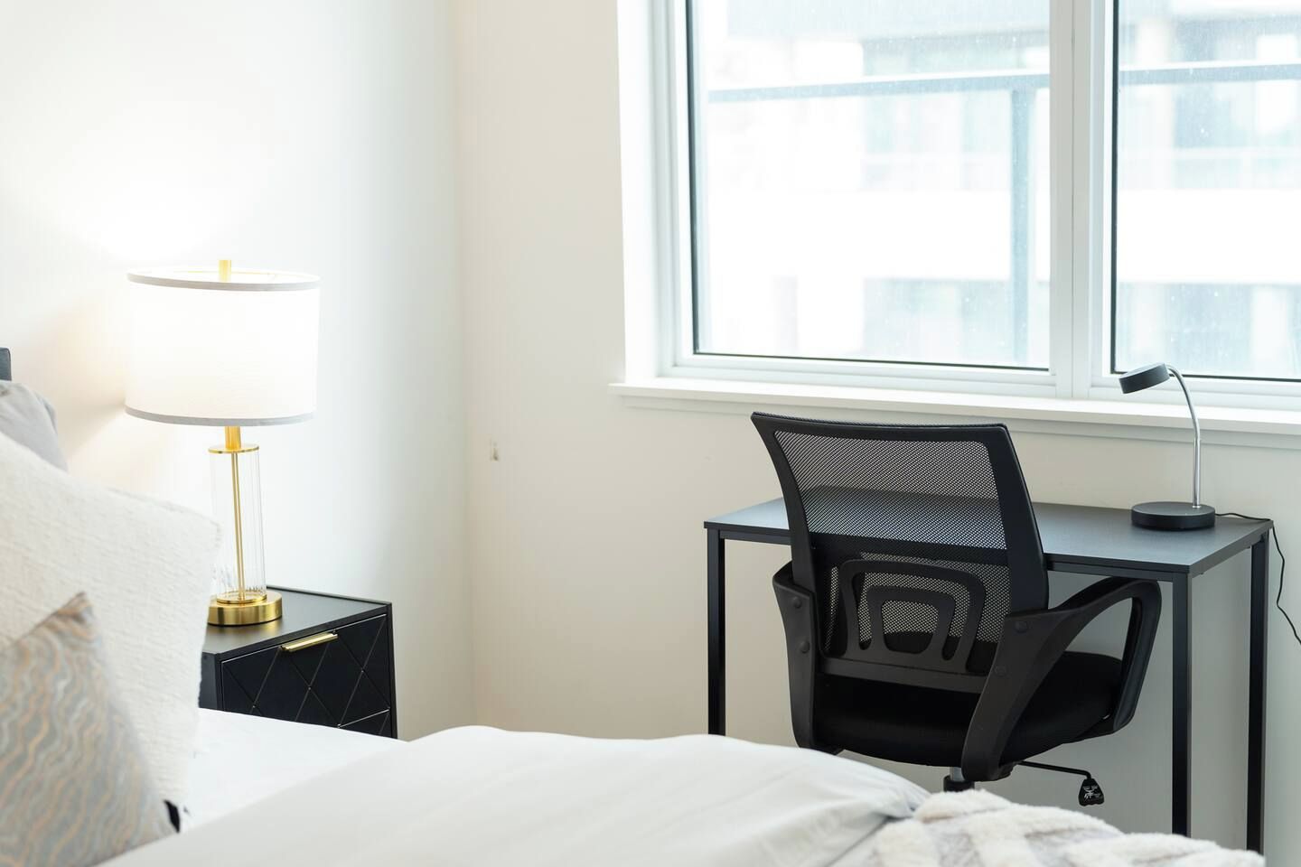 A hotel room with a bed, a desk with a chair, a lamp, and a window with a view of buildings.