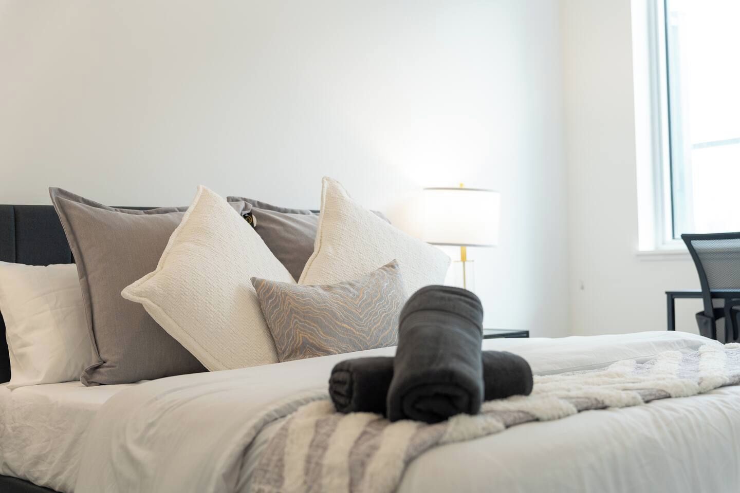 A neatly made bed with pillows and a rolled towel, a lamp, and a chair in a bright room with a window.