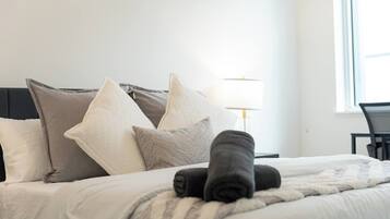 A neatly made bed with pillows and a rolled towel, a lamp, and a chair in a bright room with a window.