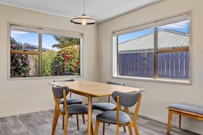 A dining area with a wooden table and chairs, large windows with views of a garden and a fence, and a hanging light fixture. - Rainbow Haven, Walk to the Lake (Taupō)