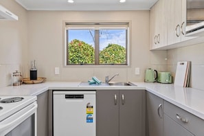 A modern kitchen with a built-in dishwasher, a window with a view of greenery, and a toaster on the counter. - Rainbow Haven, Walk to the Lake (Taupō)