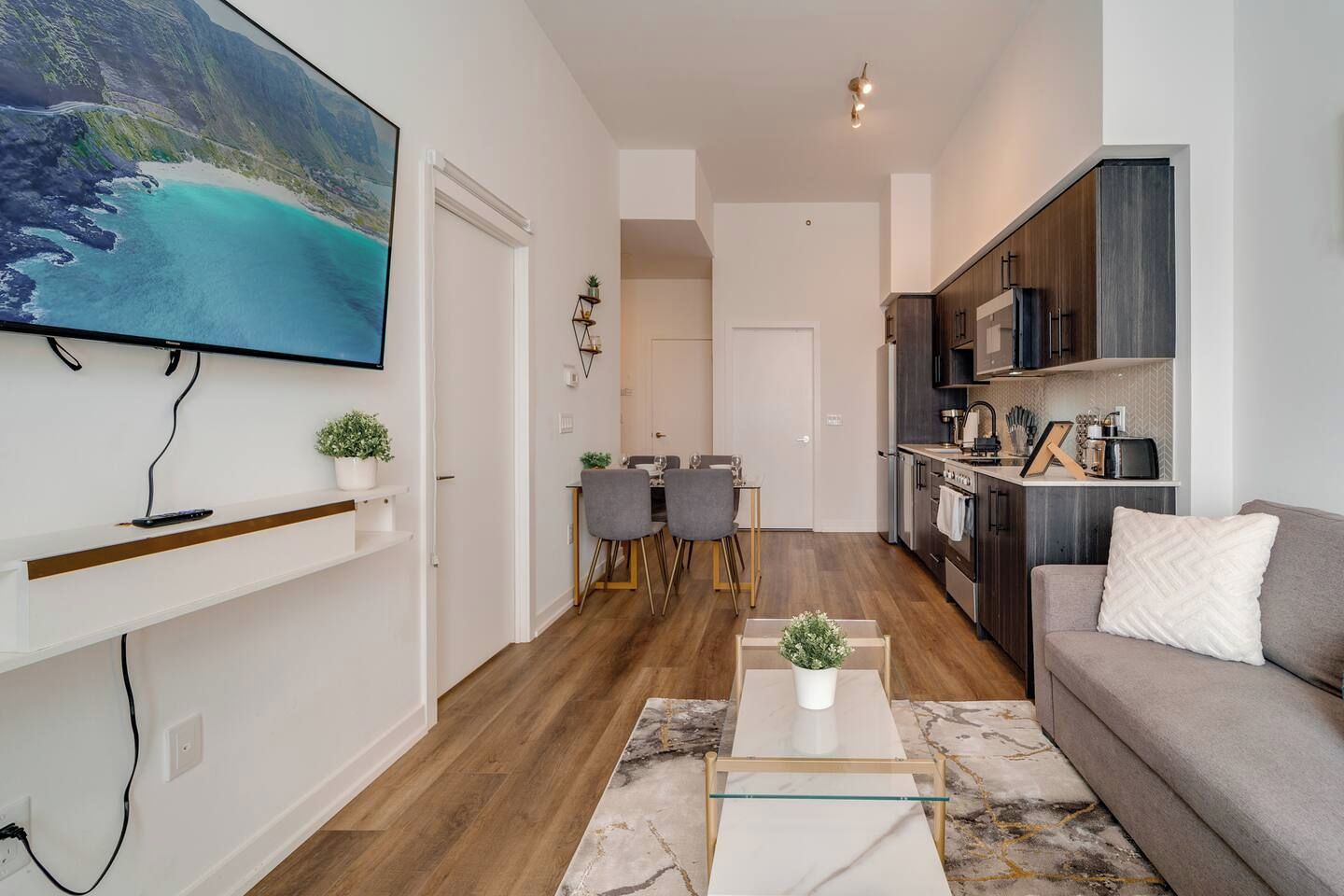 Een modern appartement met een woonkamer, eethoek en keuken. Er is een televisie aan de muur bevestigd, een eettafel met stoelen en een keukenblad met apparatuur.