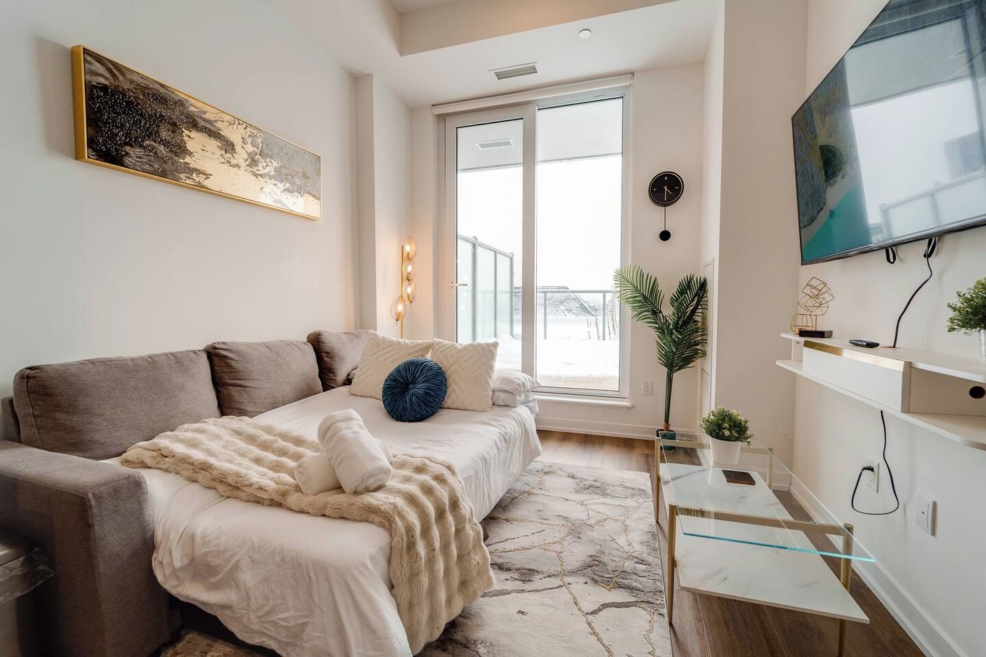A modern bedroom with a sofa bed, a flat-screen TV mounted on the wall, a bedside table with a lamp, and a large window providing a view of the outdoors.
