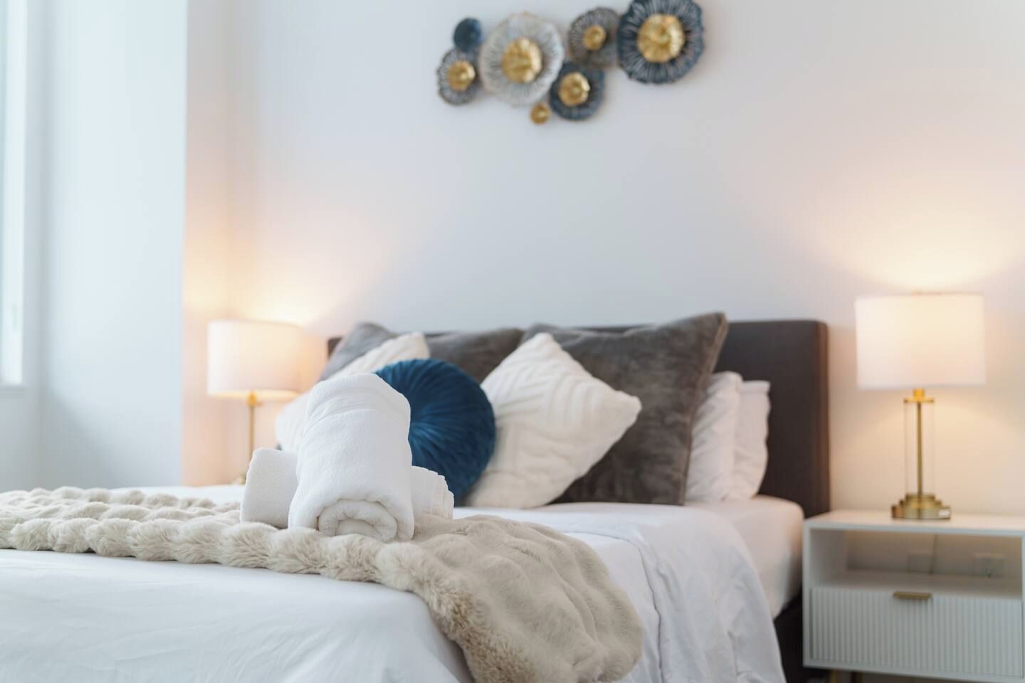 Een keurig opgemaakt bed met een zachte, witte deken, een blauw kussen en een wit kussen. Twee nachtkastjes met lampen staan aan weerszijden van het bed.