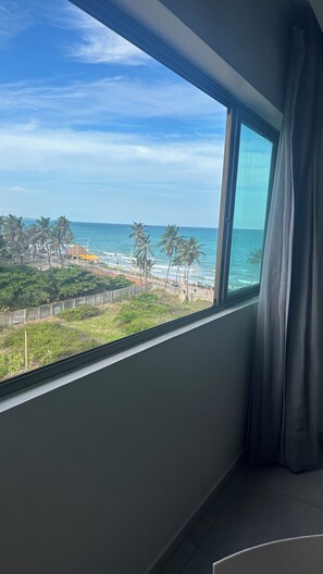 Ein Zimmer mit großem Fenster mit Blick auf einen Strand, an dem Palmen und das Meer zu sehen sind.