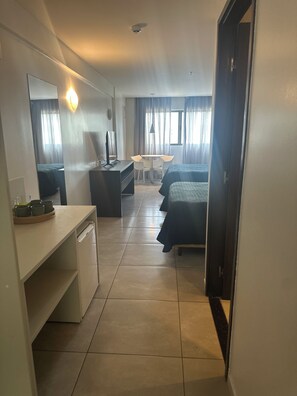 Habitación de hotel con cama, escritorio, silla, mesita, espejo y ventana con cortinas.