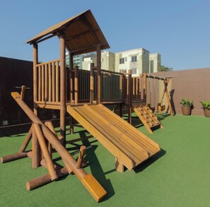 A wooden play structure with a slide and climbing elements, set on a green surface with buildings in the background. - Apartment in luxury boutique hotel Ritz Suites Beira Mar, with pool, gym and (Maceió)