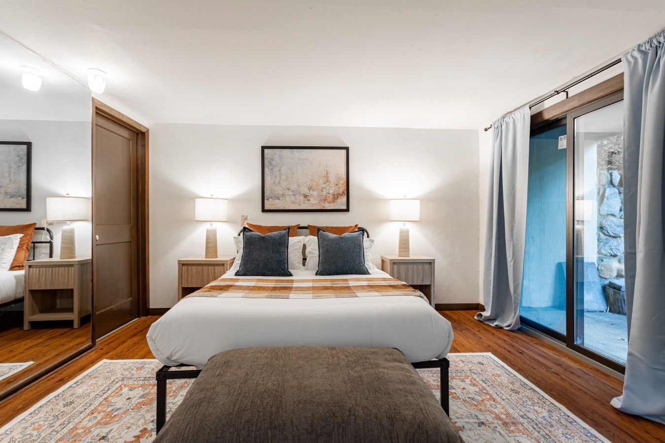 Ein Hotelzimmer mit einem großen Bett, zwei Nachttischen, einer Bank, einem großen Fenster mit Vorhängen und einem Gemälde an der Wand.