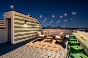 A rooftop lounge with a green-striped wall, outdoor seating, string lights, and a neon sign. - Nashville rooftop retreat with fire pit, garage, sleeps 12 near Geodis Park (Nashville)