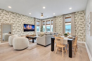 A living room with a sofa, a dining table with chairs, a TV mounted on the wall, and a large window with curtains. - rooftop deck, fire pit & skyline views near Geodis Park | sleeps 12 (Nashville)