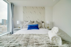 A hotel room with a large bed, two bedside tables, a window with curtains, and a city view. - Modern Condo in Central KW w/ Gym (Kitchener)