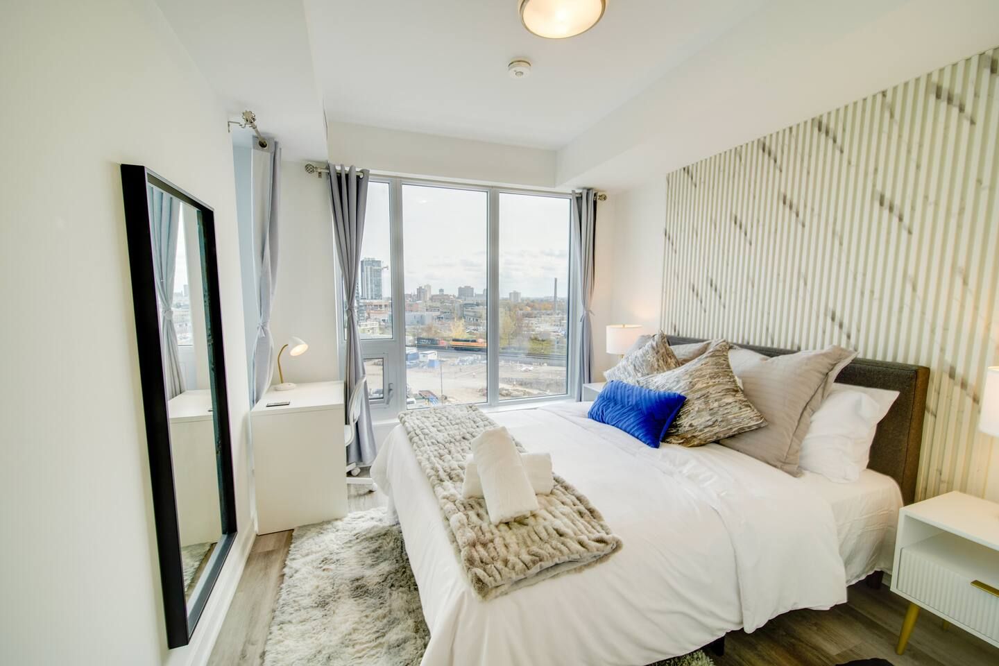 A modern hotel room with a large bed, a nightstand, a mirror, and a view of the city through large windows.