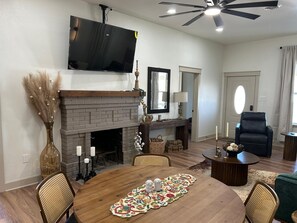 Un salon avec une cheminée, un téléviseur fixé au-dessus, une table à manger avec des chaises et un ventilateur de plafond.