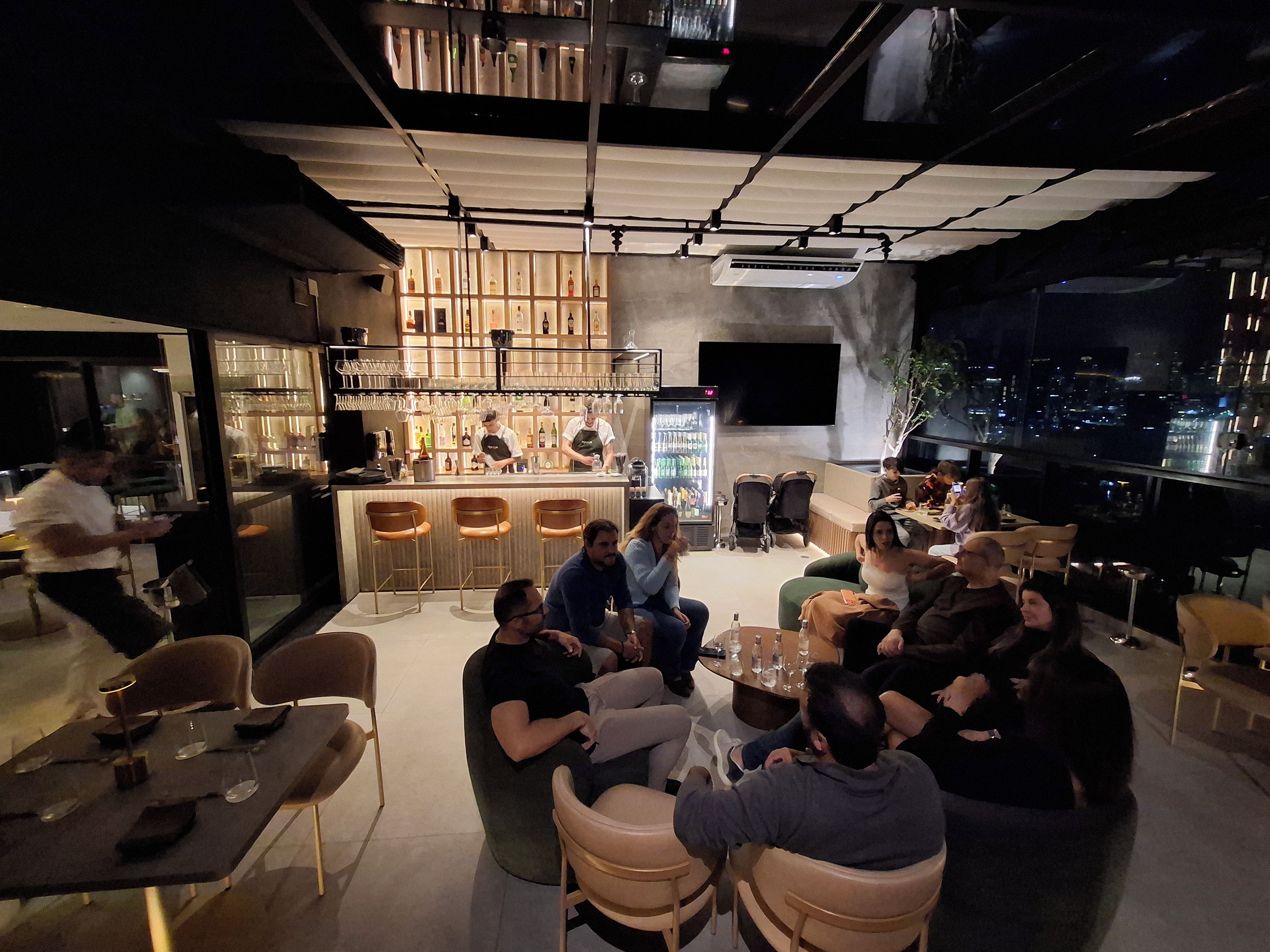 Un área de bar moderna con disposición de asientos para los huéspedes, un barman en la barra y una vista panorámica de la ciudad al fondo.