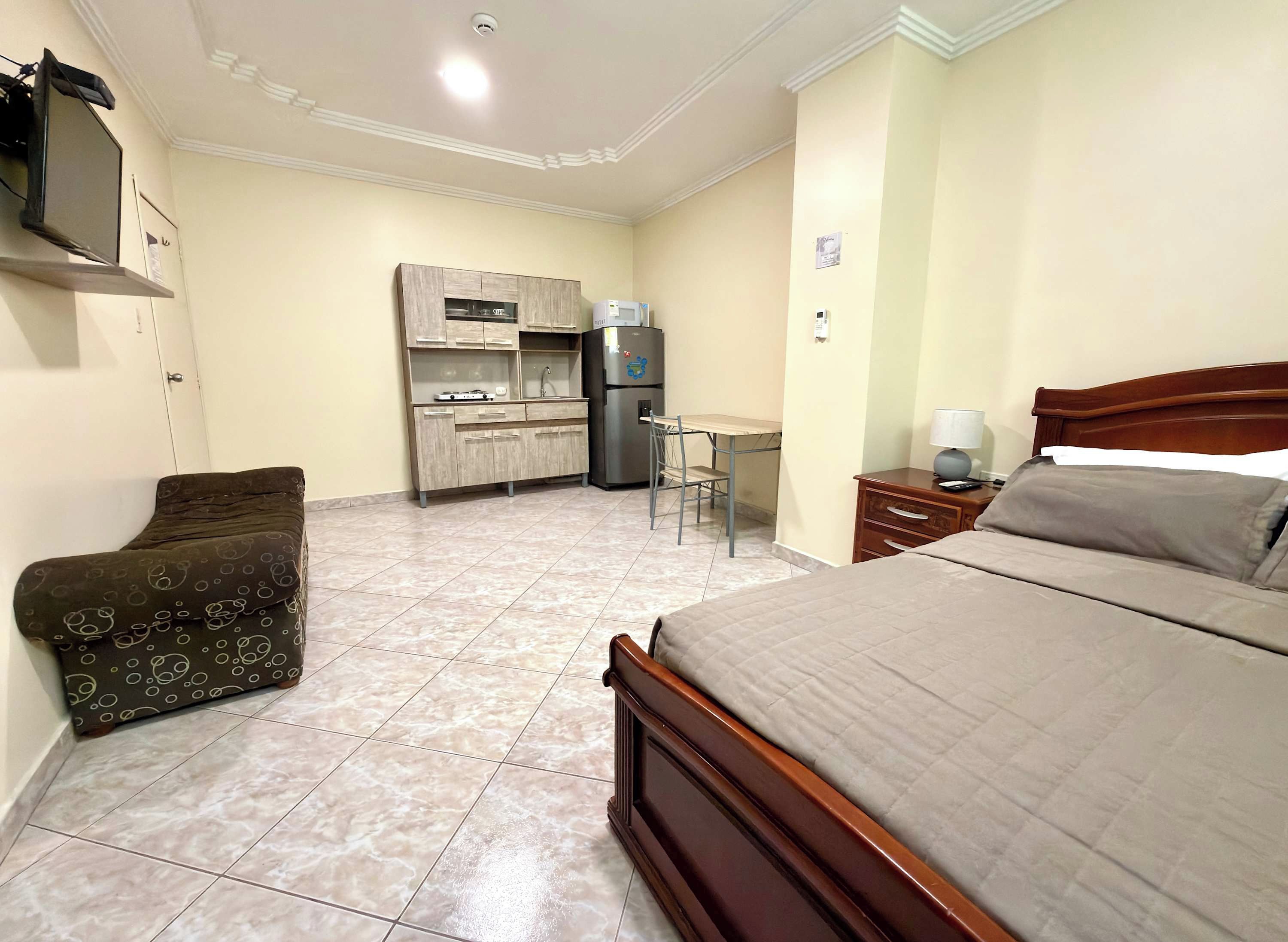 A hotel room with a bed, a desk, a chair, a kitchenette, and a TV.