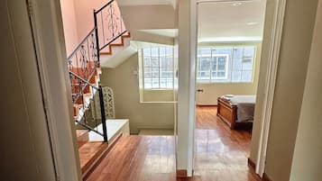 A hallway with wooden flooring, a staircase with metal railings, and a door leading to a room with a bed and a window.