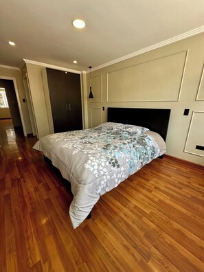 Un dormitorio con una cama grande, suelo de madera, cabecera oscura y una lámpara en el techo.