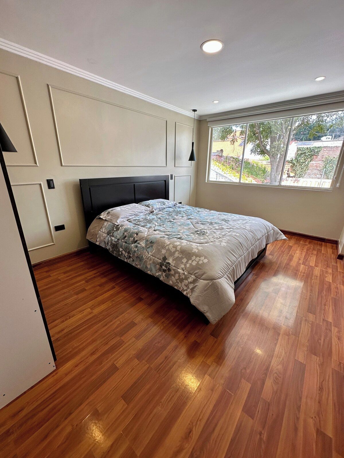 A bedroom with a bed, wooden flooring, a window with a view, and recessed ceiling lighting.