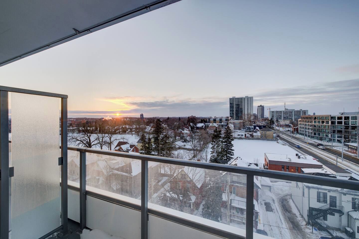 A balcony with a view of a cityscape during sunset, featuring buildings, a snow-covered ground, and a clear sky.