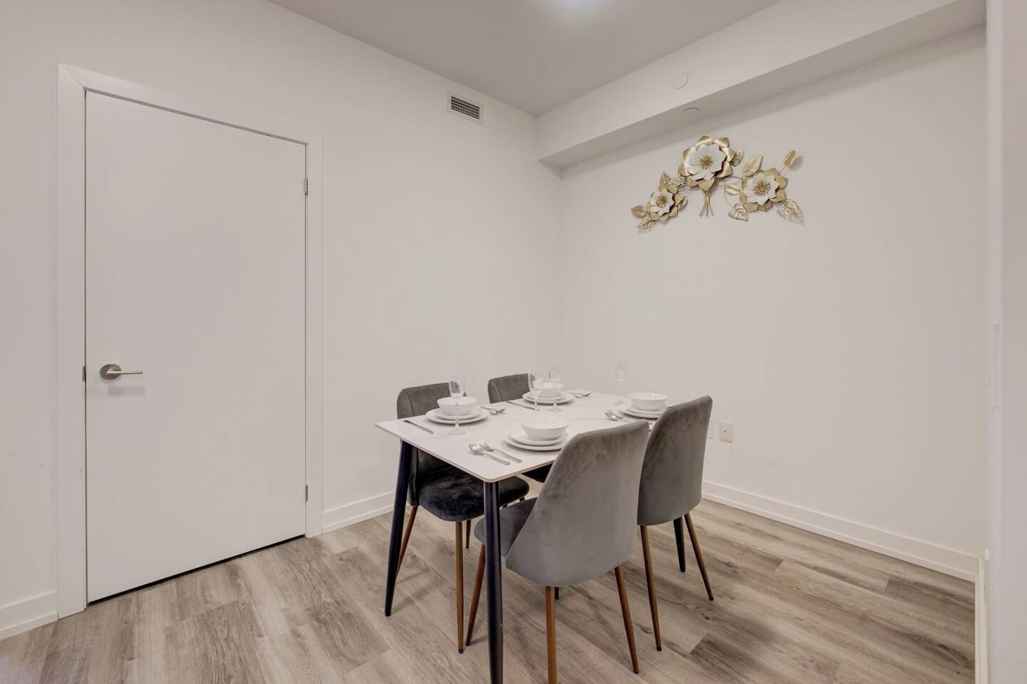 A dining area with a white table set for four, grey chairs, and a decorative wall piece.