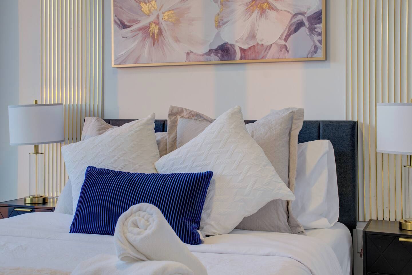 Een opgemaakt bed met witte en blauwe kussens, een opgerolde witte handdoek en een schilderij van bloemen aan de muur.