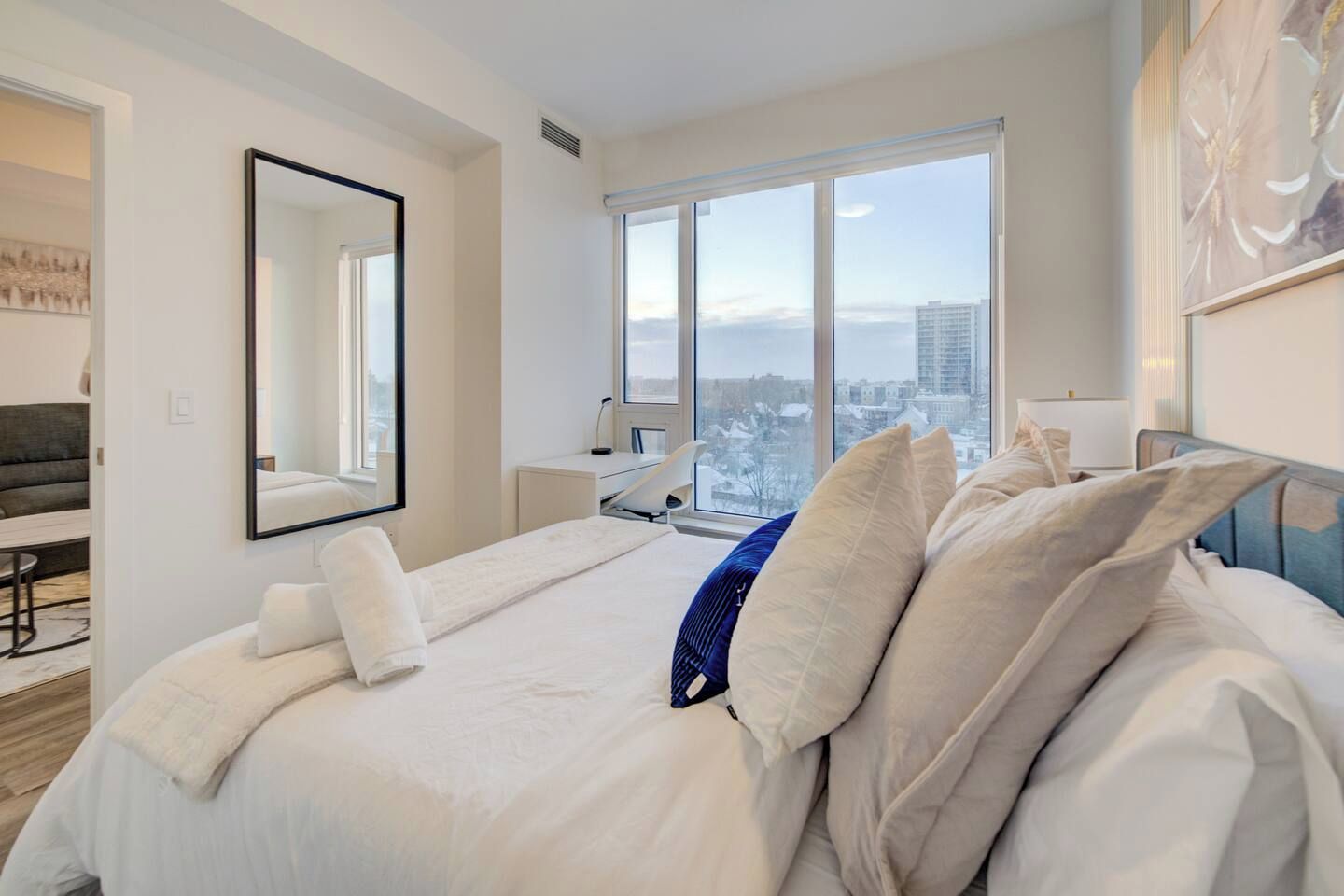A modern hotel room with a large bed, a desk, a chair, and a view of the city.