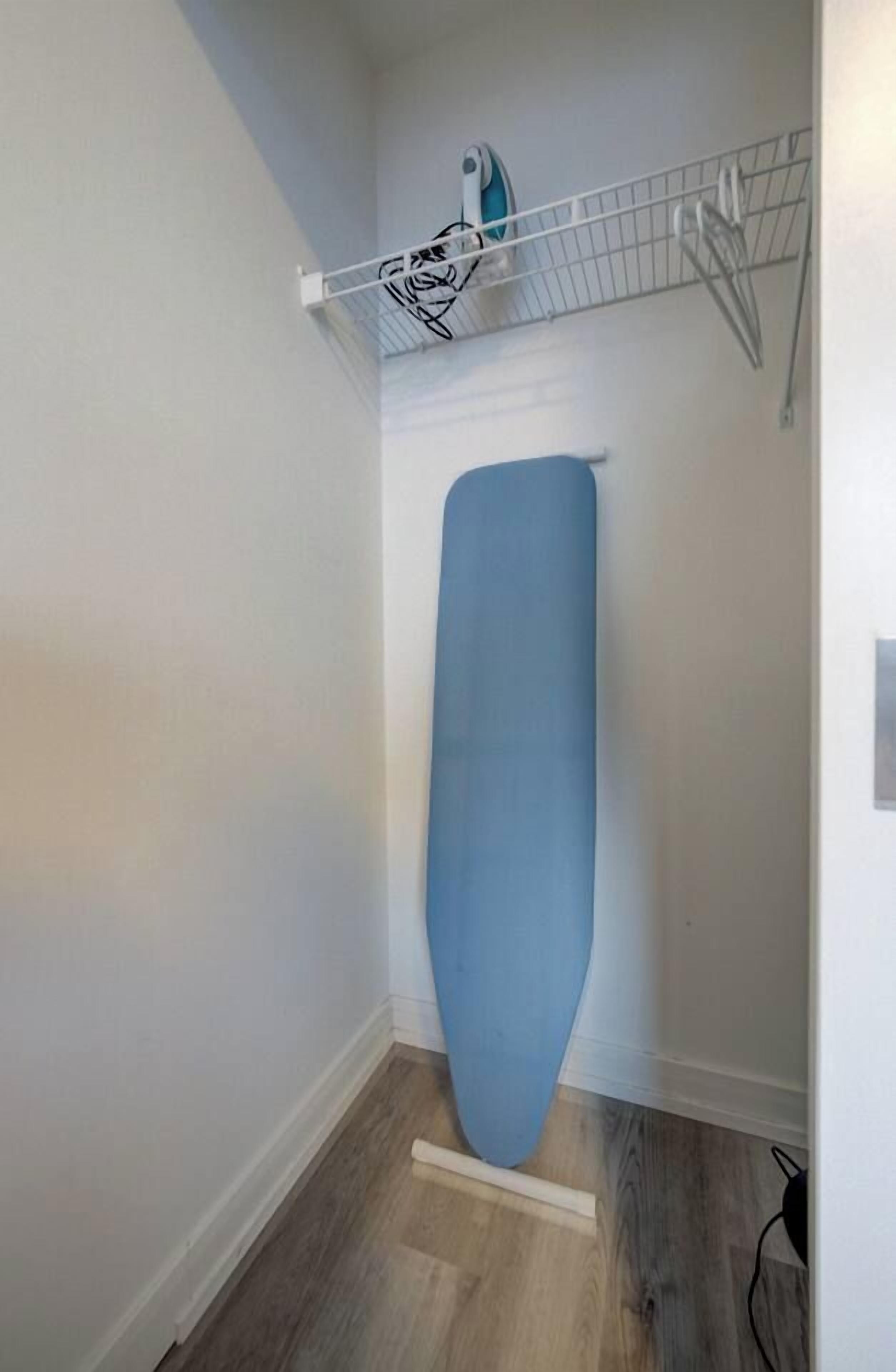 A room with a wooden floor, a white wall, and a metal shelf with a blue ironing board.