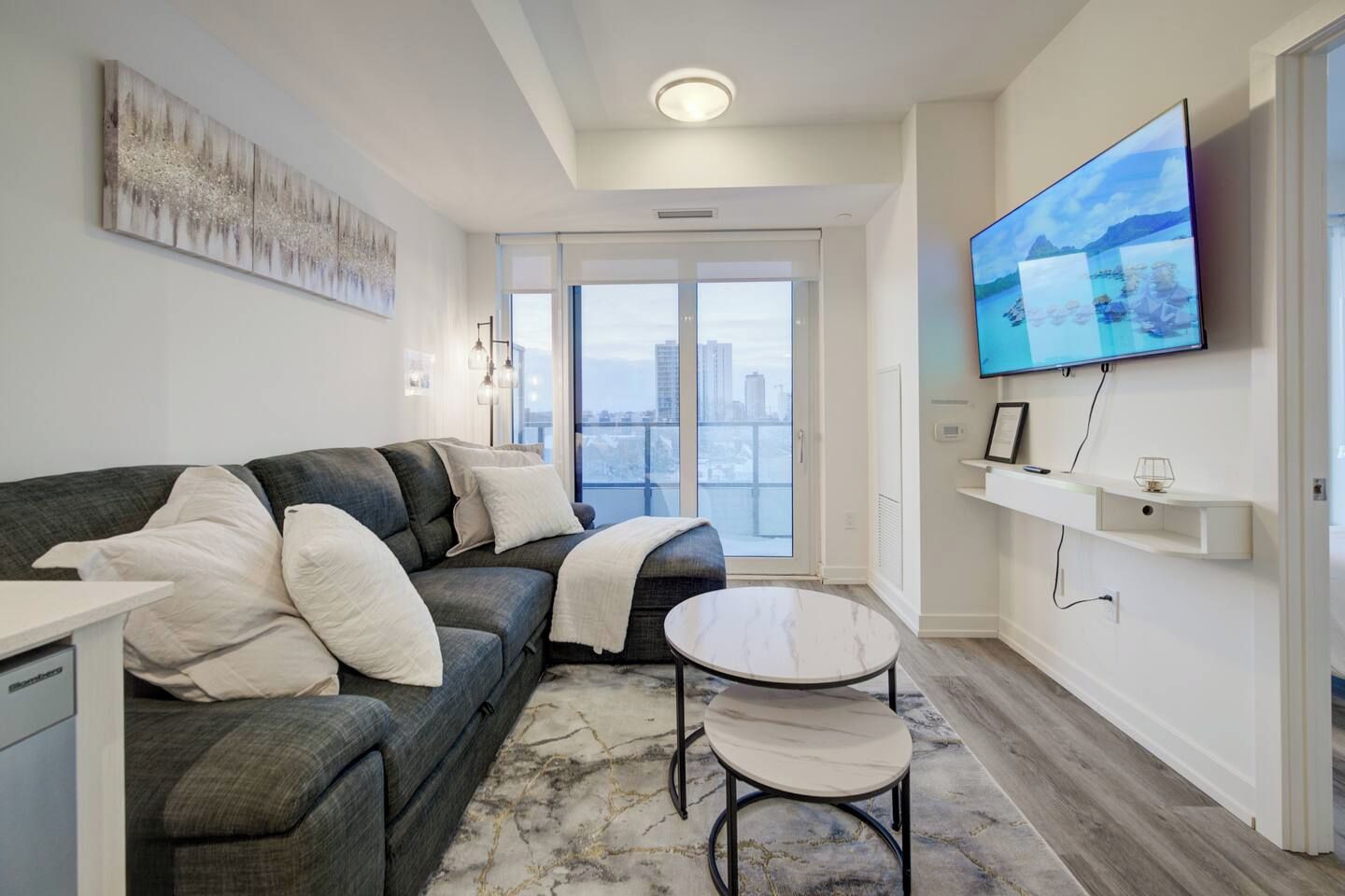 Un salon moderne avec un canapé gris, une petite table ronde et un téléviseur à écran plat fixé au mur.