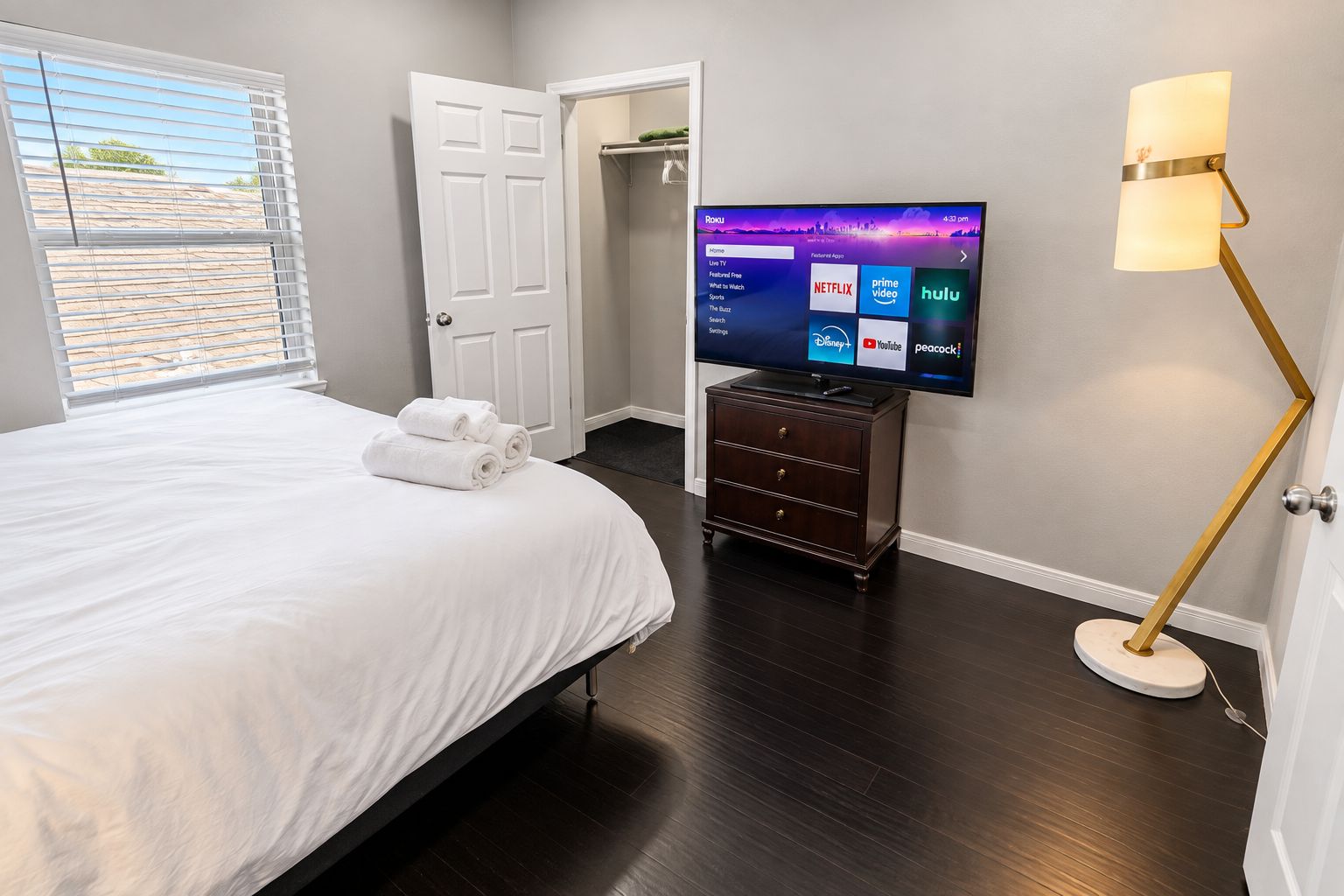 A hotel room with a bed, a flat-screen TV mounted on the wall, a wooden dresser, and a floor lamp.
