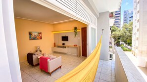 A balcony with a yellow hammock, a sofa, a TV, and a painting on the wall. - Apto c Varanda e Vaga pr x a Praia Porto da Barra (Salvador)