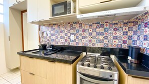 A compact kitchen with a gas stove, a microwave, and a sink. The backsplash is tiled with a colorful pattern. - Apto c Varanda e Vaga pr x a Praia Porto da Barra (Salvador)
