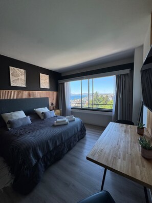 A modern bedroom with a large bed, a wooden table, and a view of a cityscape. - Vista al lago a pasos del Centro Cívico (San Carlos de Bariloche)