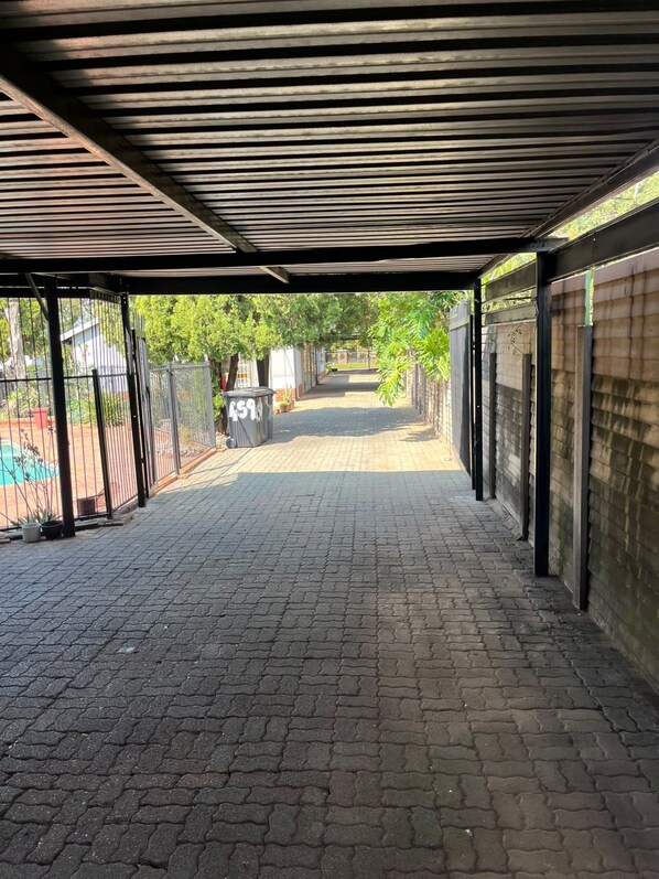 Un espacio al aire libre techado con un sendero pavimentado, cerca metálica y vistas a una zona residencial a lo lejos.