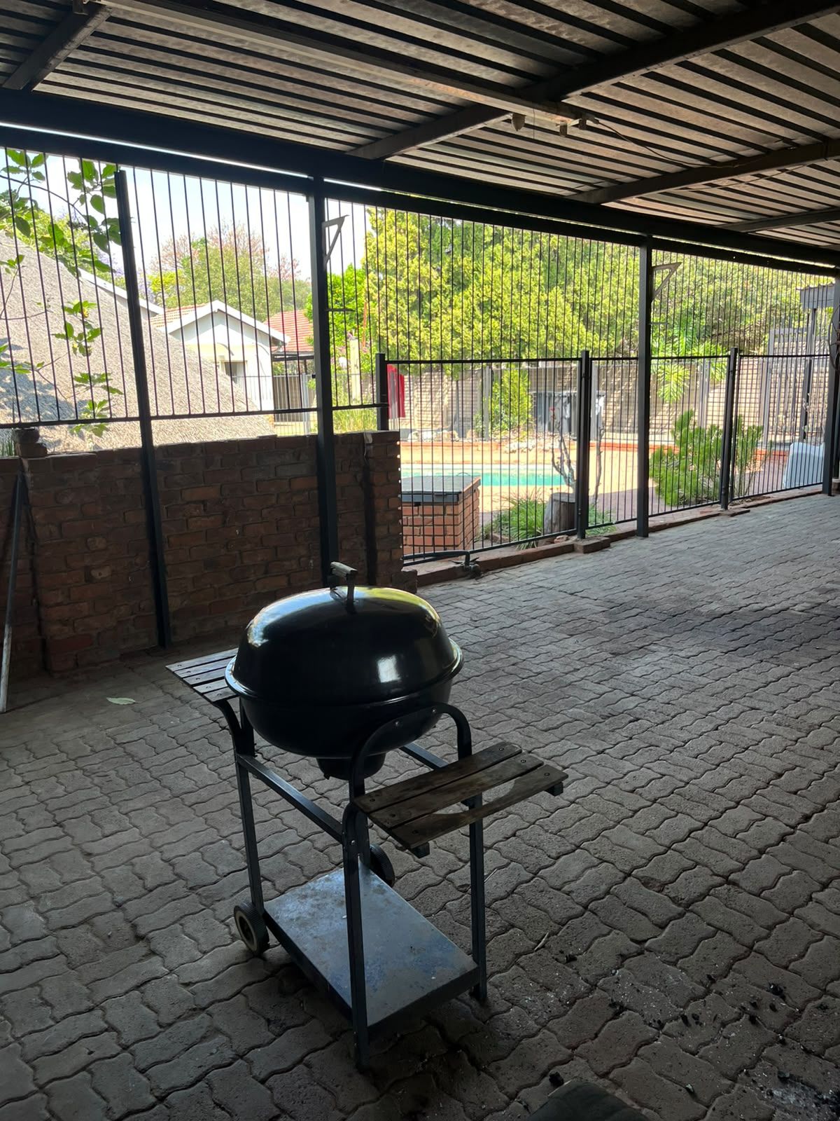 A covered outdoor area with a barbecue grill on a cart, brick walls, and a view of a swimming pool and greenery.