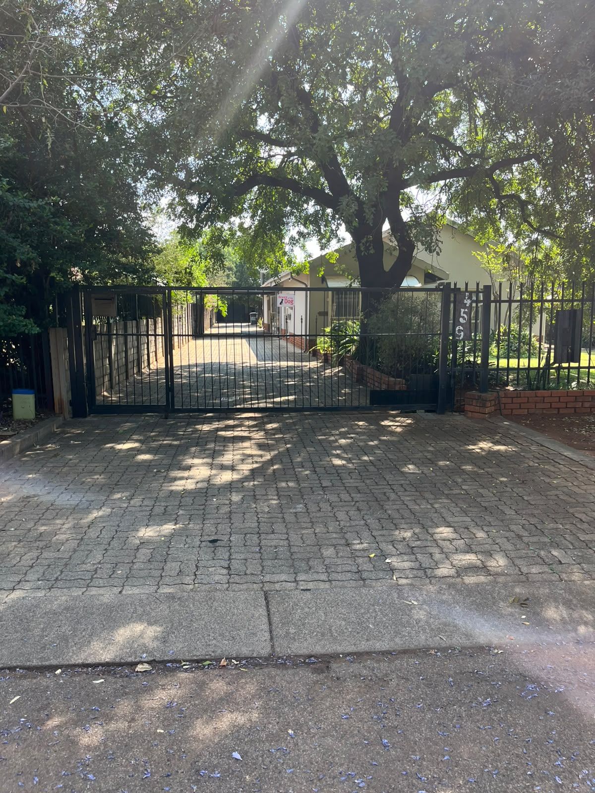 A gated driveway leading to a building with a paved entrance and a tree providing shade.
