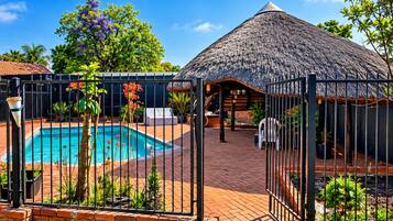 A swimming pool with a fenced area, surrounded by greenery and a thatched-roof structure.