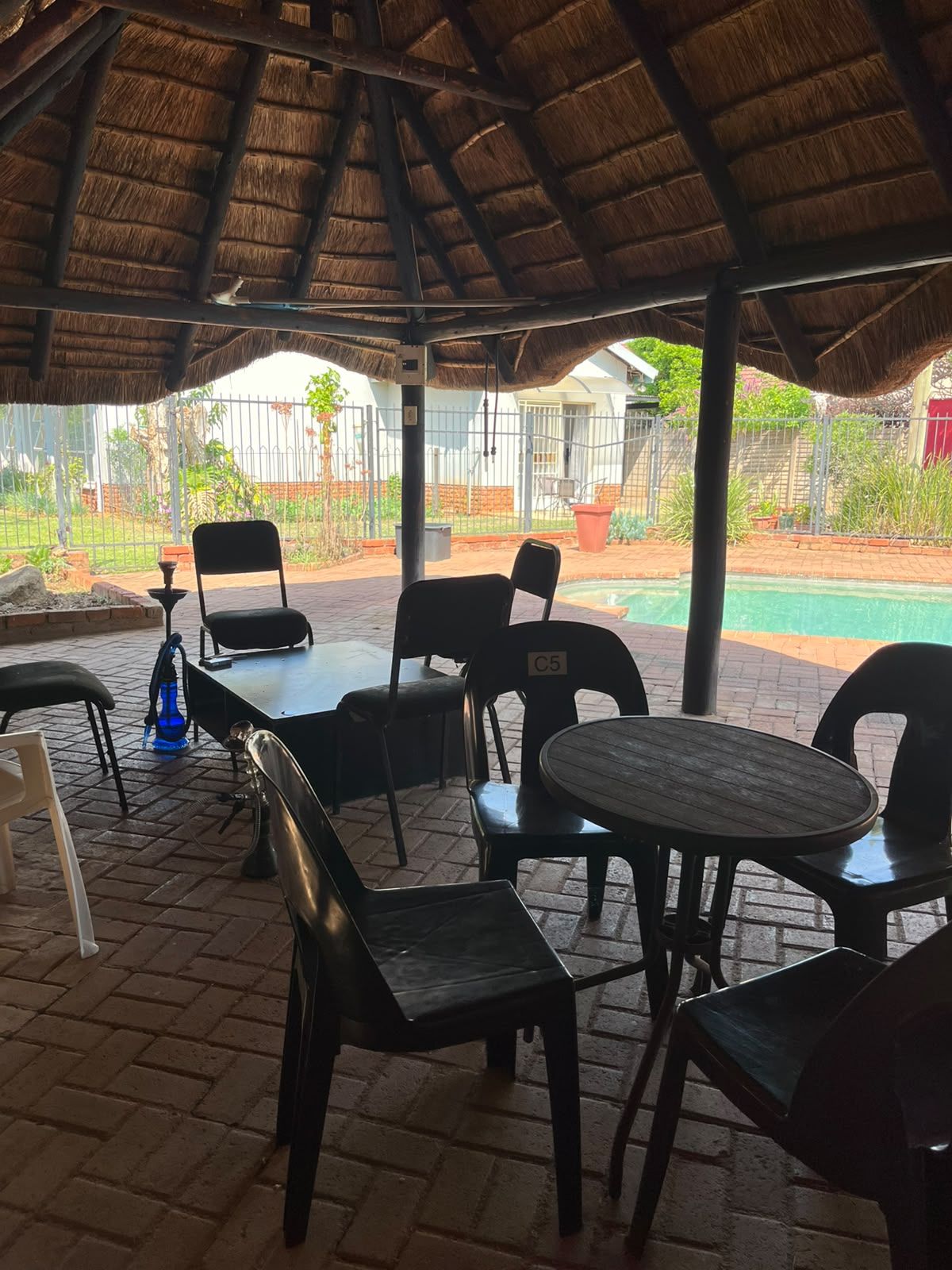 An outdoor seating area with tables and chairs, a thatched roof, and a view of a swimming pool and greenery.