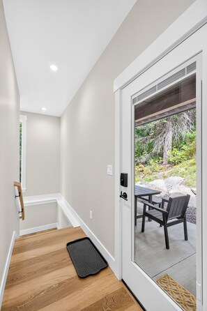 A modern interior with a wooden staircase, a black doormat, and a sliding glass door leading to an outdoor patio with a table and chairs. - Private Hot Spring | Views | River | Brundage (New Meadows)
