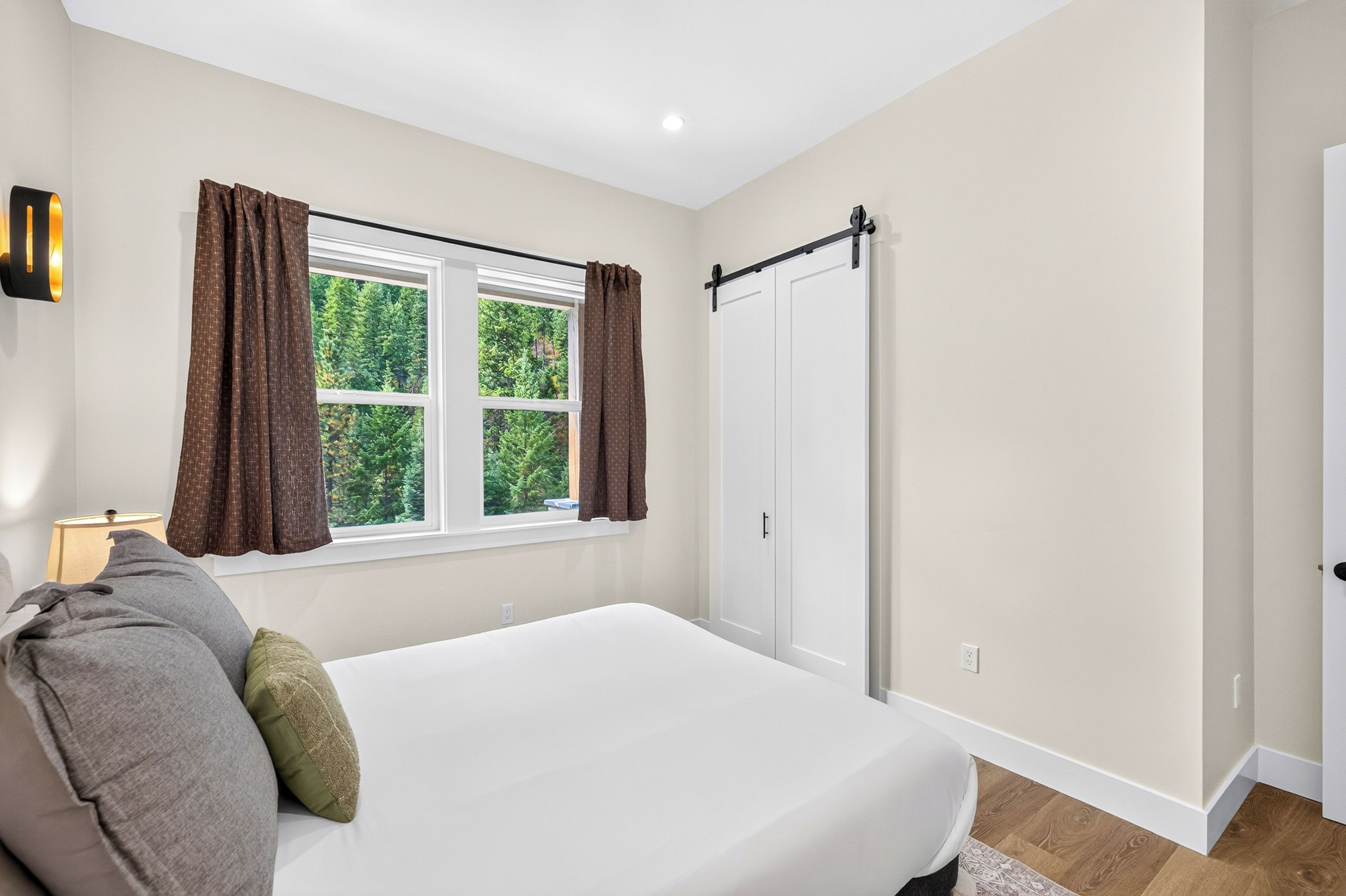 A bedroom with a bed, a window with curtains, a wall-mounted lamp, and a sliding door.