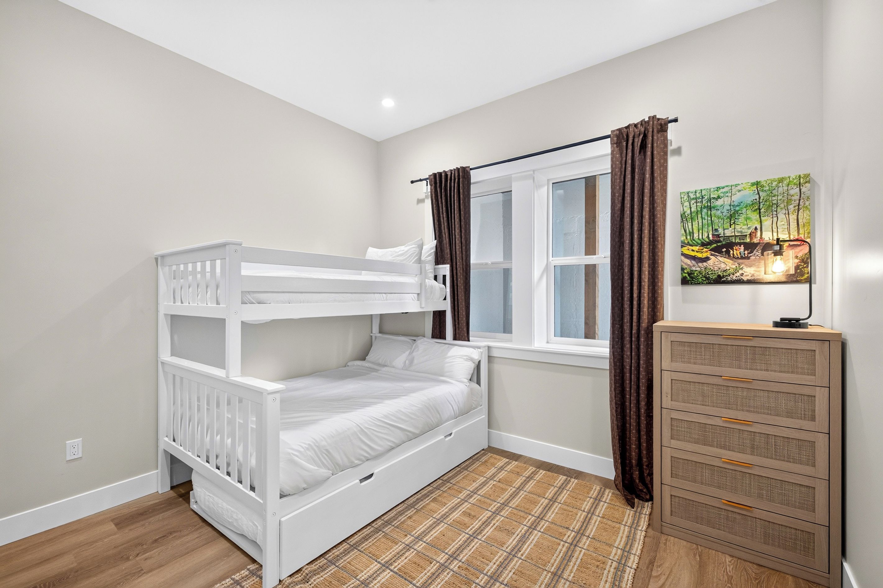 A child’s room with a bunk bed, a window with curtains, a painting on the wall, and a dresser with drawers.