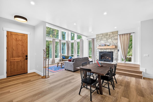 Una sala de estar amplia con chimenea, ventanas grandes y una zona de comedor con mesa y sillas de madera.