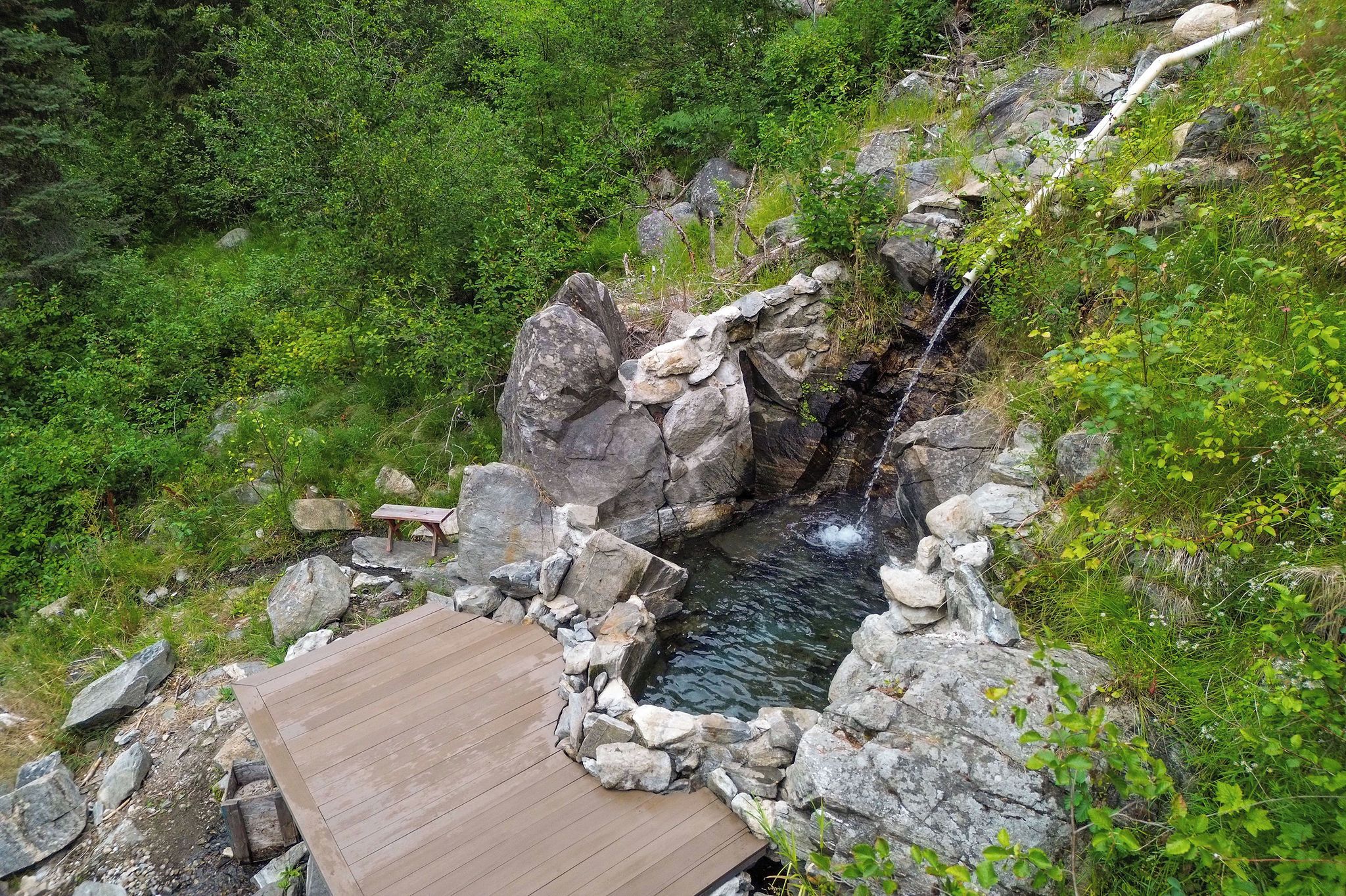 Une terrasse en bois surplombant une source d’eau chaude naturelle, avec un mur de pierres et des rochers.