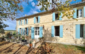 Ein Gebäude aus Stein mit blauen Fensterläden, ein Garten mit Kübelpflanzen und einem klaren Himmel.