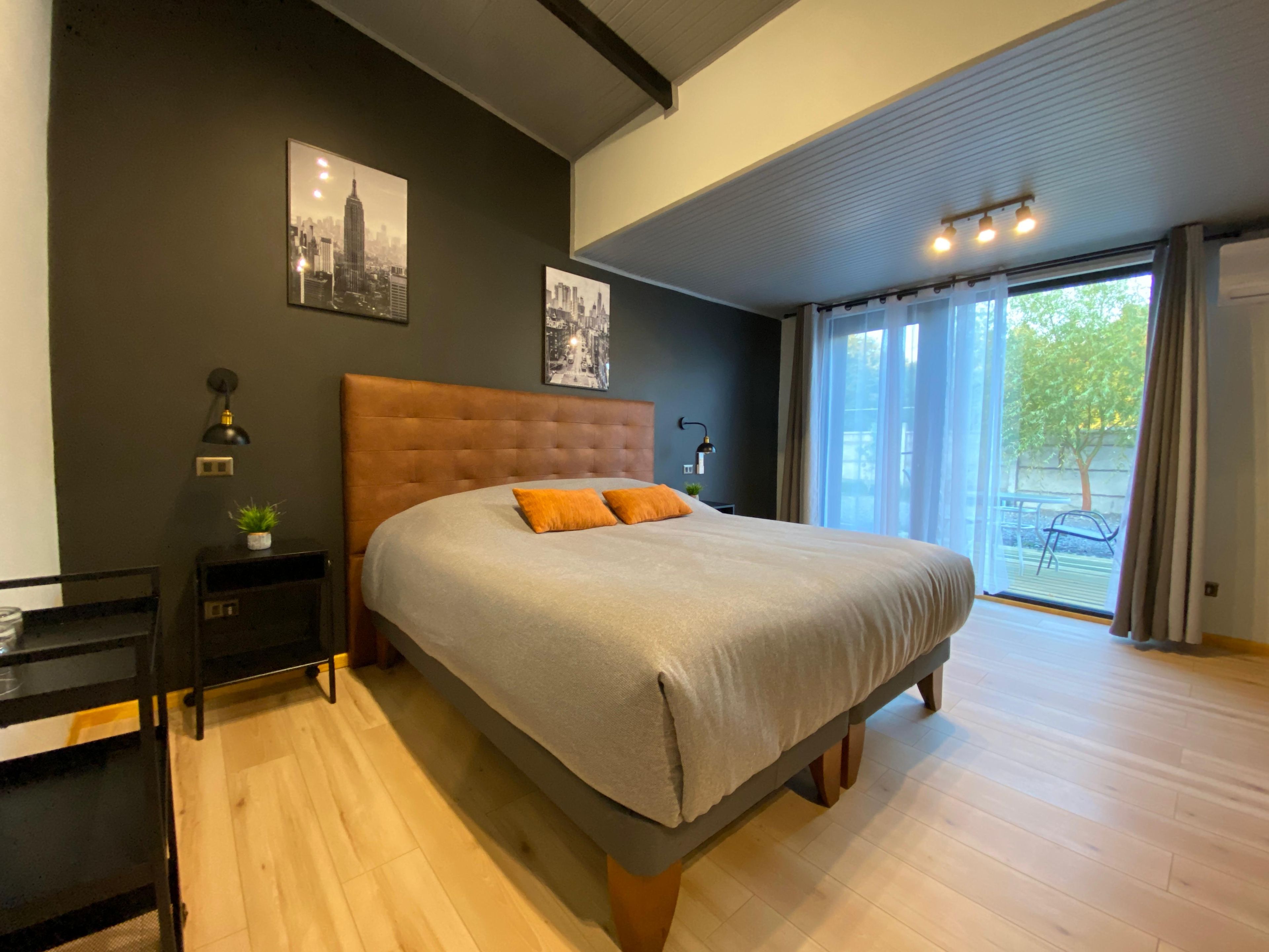 A bedroom with a large bed, a nightstand, a wall-mounted lamp, and two framed pictures.