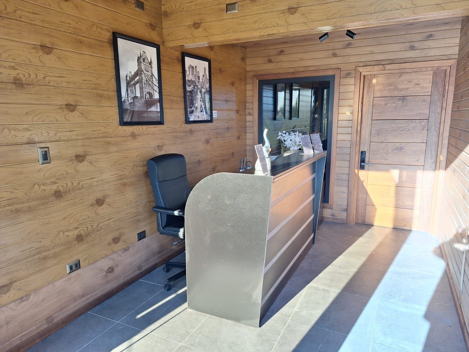 Un área de recepción con paneles de madera, un mostrador de recepción, una silla de oficina negra y cuadros enmarcados en la pared.