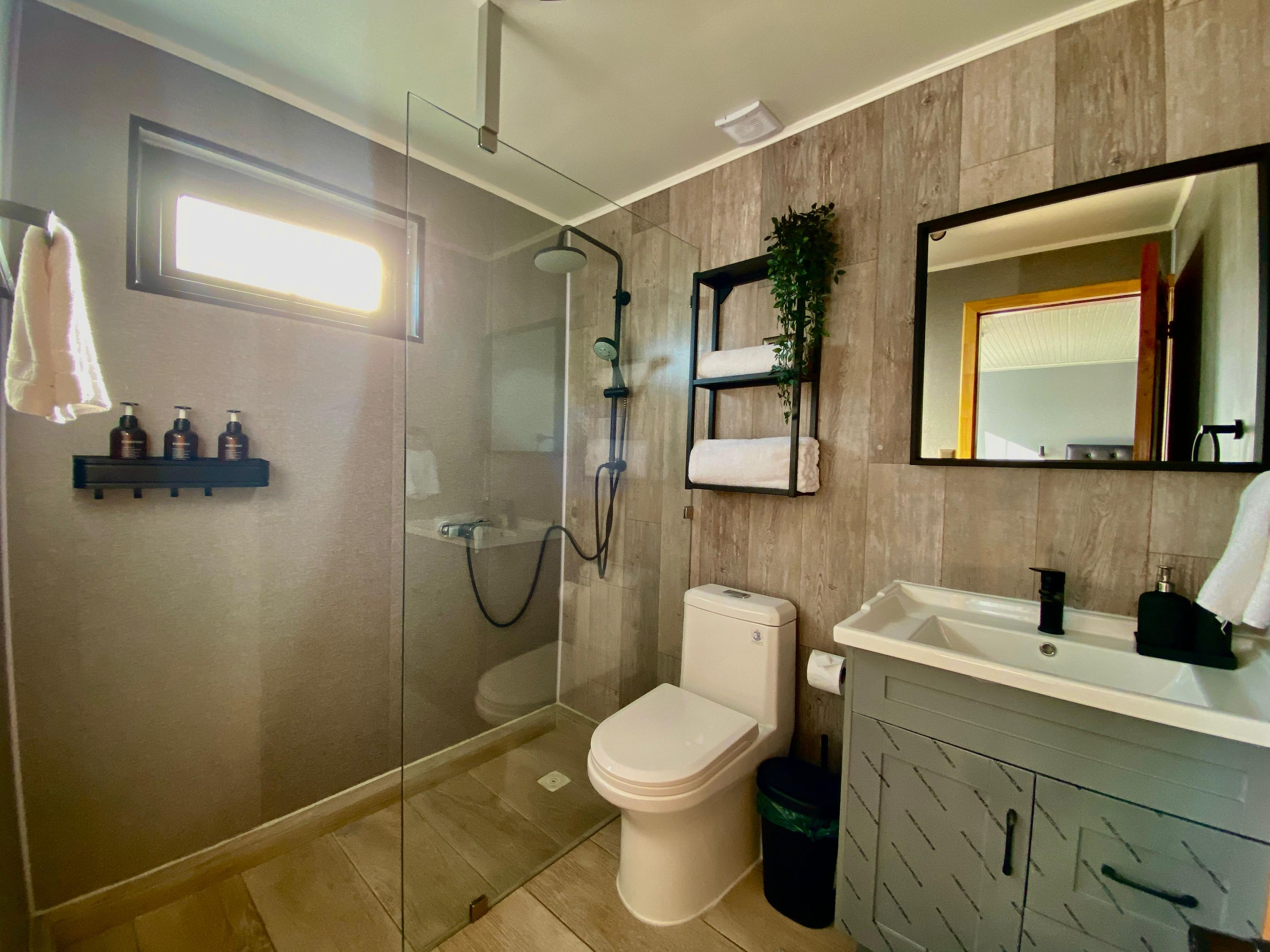 A modern bathroom with a glass-enclosed shower, a toilet, a sink with a mirror, and a shelf with toiletries.